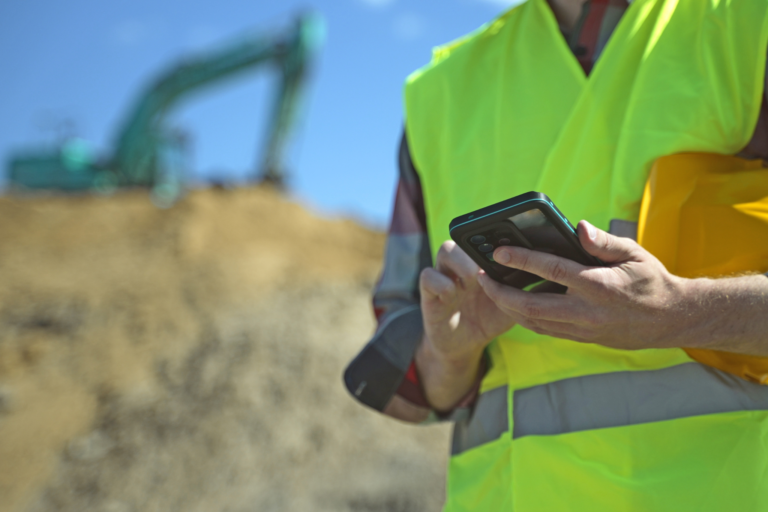 Construction worker holding phone researching blue cscs cards in green illuminous high vis jacket.