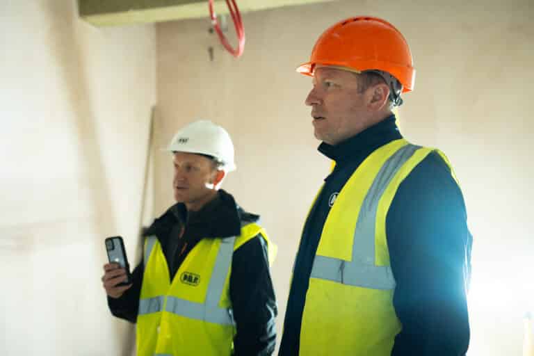 Two construction workers examining a wall and on their phone searching how to manage a worker with an incorrect or expired CSCS card.