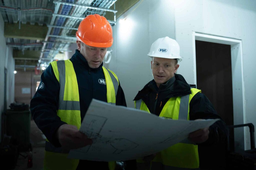 Two construction workers in DMR Training hi-vis and hard hats reviewing site drawings during an on-site NVQ Level 2 assessment