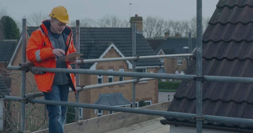Site supervisor in hi-vis and hard hat checking progress on a tablet from scaffolding on a UK housing site